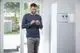 Mann steht in modernem Raum und schaut auf sein Smartphone. Im Hintergrund ist eine weiße Heizungswärmepumpe von Vaillant zu sehen, zusammen mit Fenstern und einer grünen Außenansicht.